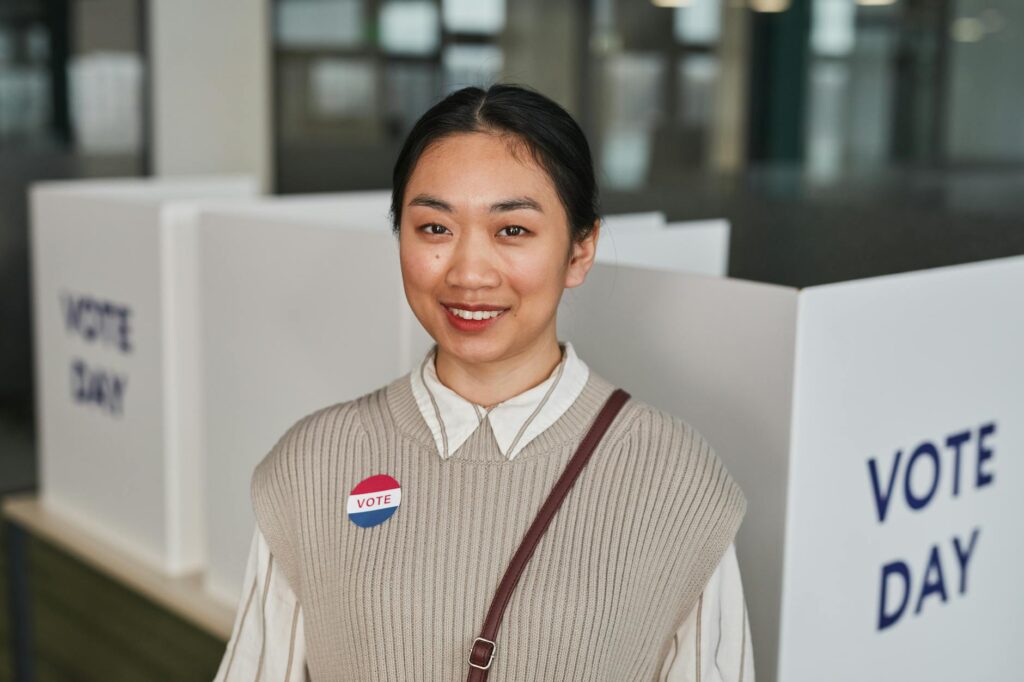 Asian woman smiling at polling station with 'Vote' badge on Voting Day.