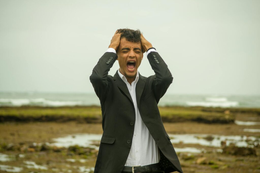 A man in a suit screams in frustration while standing on a windy beach. Emotional stress concept.