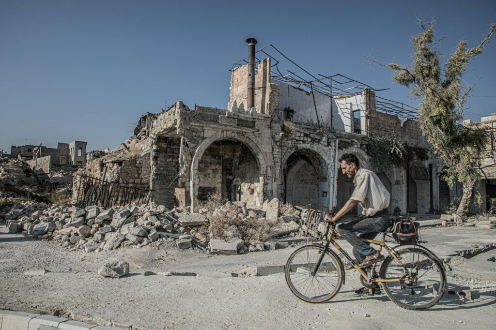 A man rides a bicycle past the ruins of Aleppo, symbolizing resilience amidst destruction.
