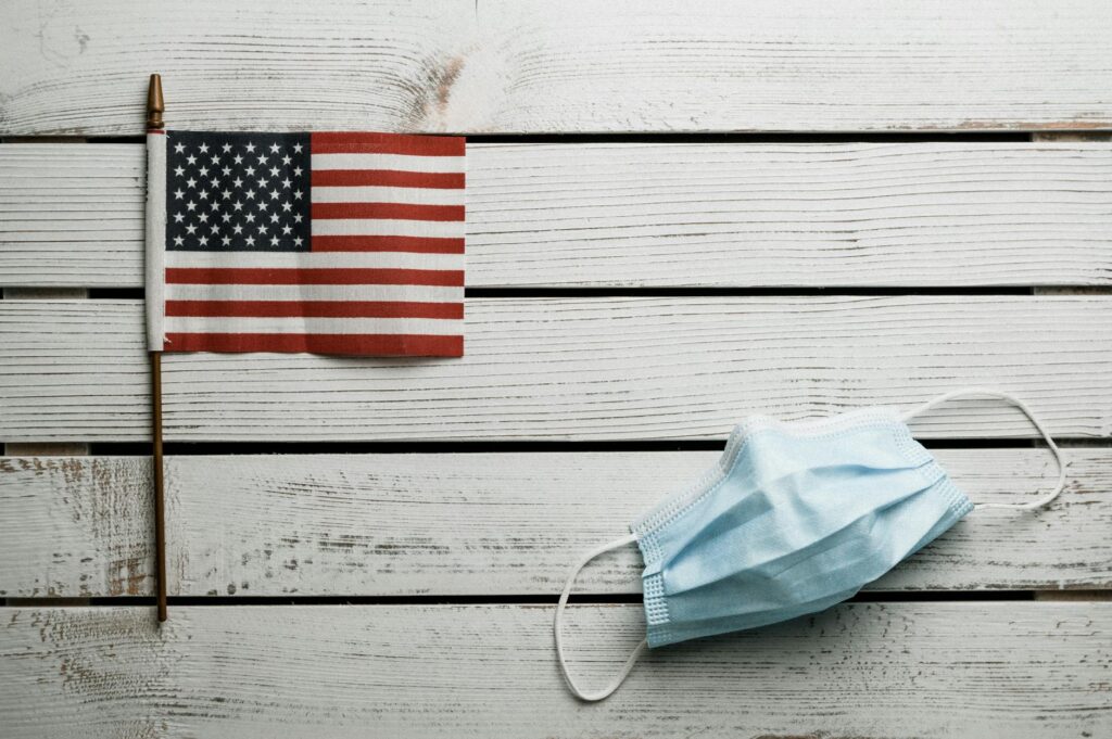 Top view of small Flag of United States and light blue fabric disposable face mask on white wooden wall