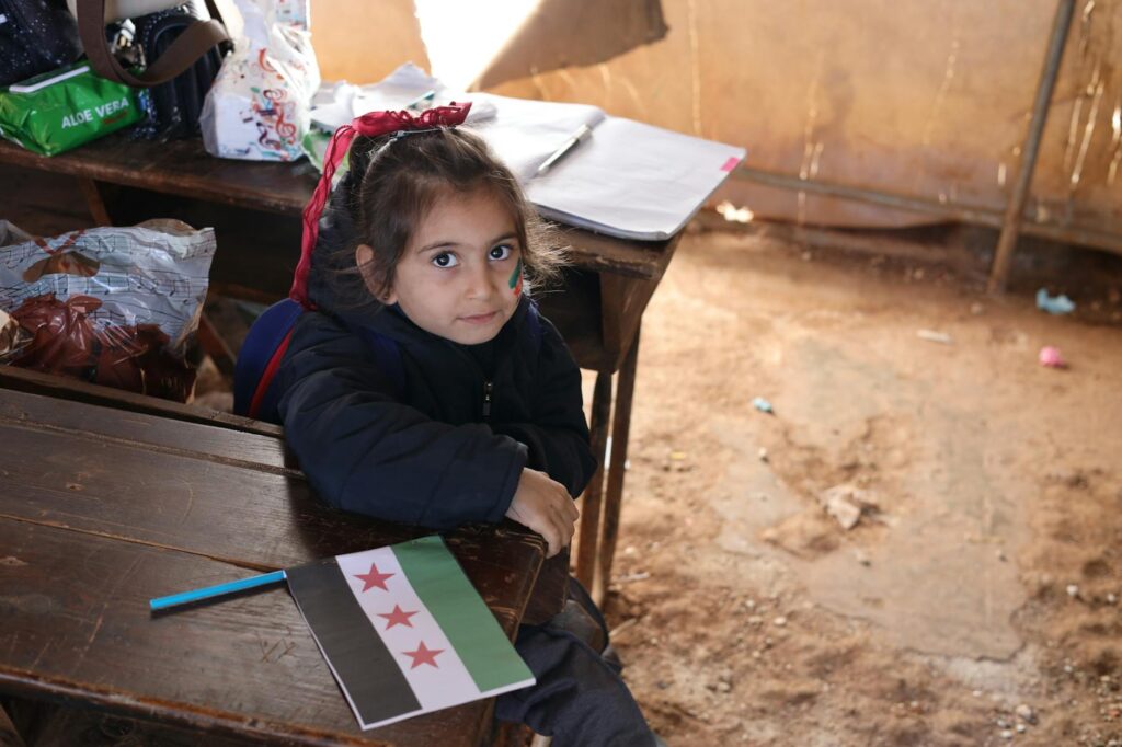 A young girl in a Syrian classroom holding a flag and smiling.