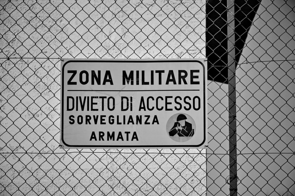Black and white photo of a restricted military zone sign behind a metal fence.