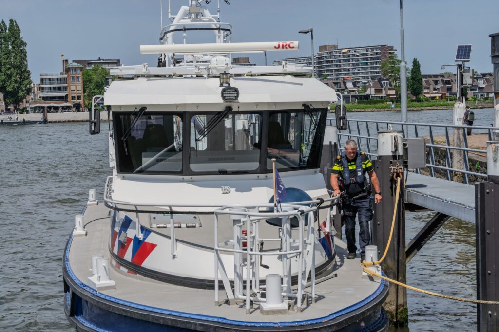 Police boat with officer docked at an urban waterfront, featuring modern buildings in the background.