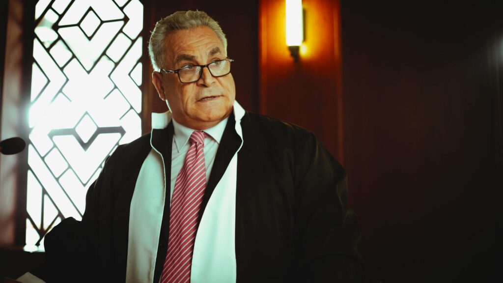 A male judge in a courtroom in Baghdad wearing a robe and tie, in a contemplative pose.