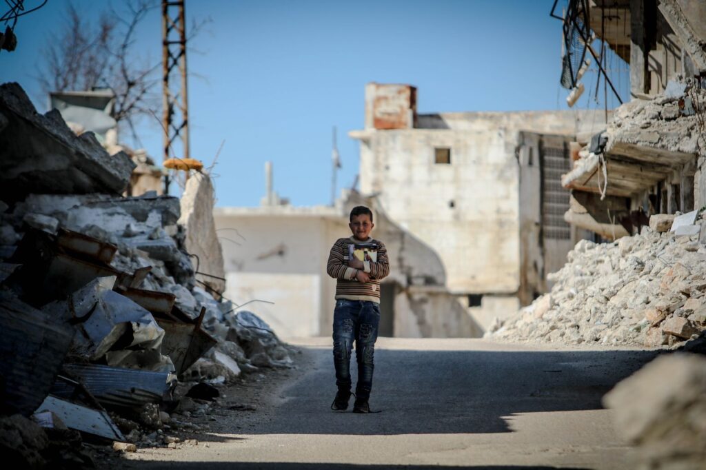 A young boy stands amidst the rubble of a war-torn area in Idlib, Syria, showcasing resilience.
