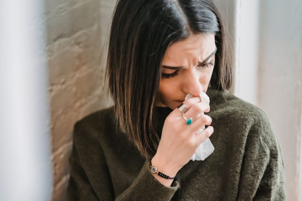 Despaired female with dark hair in casual clothes standing near wall with tissue in hand in light room at home