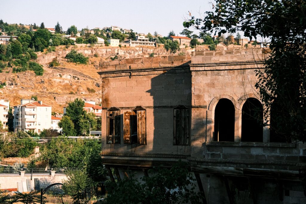 Sunlit historic stone building with arched windows amid lush landscape and hills, representing architectural heritage.