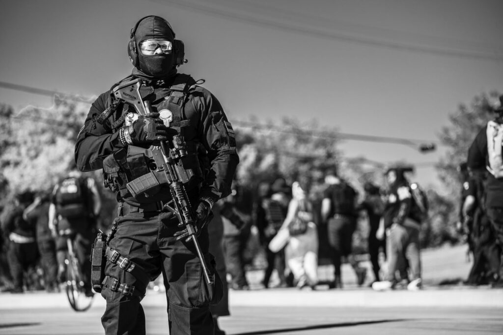 Monochrome image of a soldier in uniform with a rifle standing on a street amidst a crowd, conveying military presence.