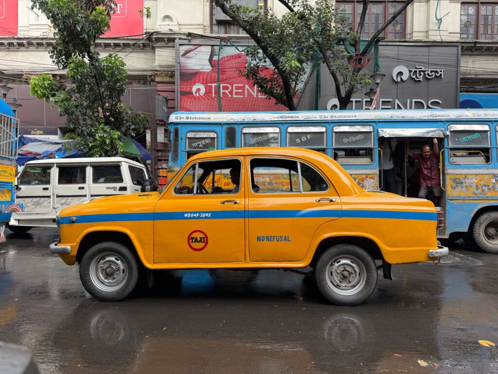 Iconic yellow taxi on a rainy Kolkata street with vintage buses and local signage.