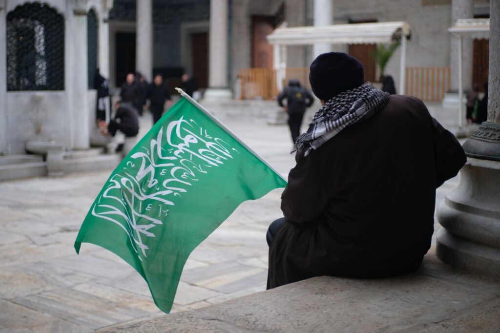 A man sits holding a green flag with Arabic script on a city street.