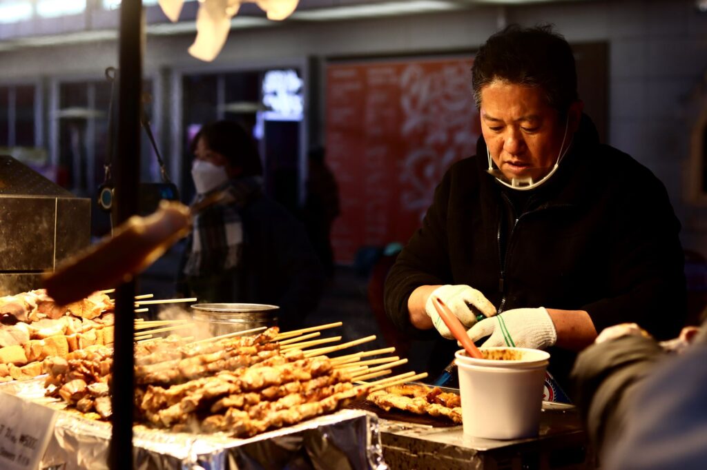 Asian street vendor cooking skewered food at a bustling market during the evening.