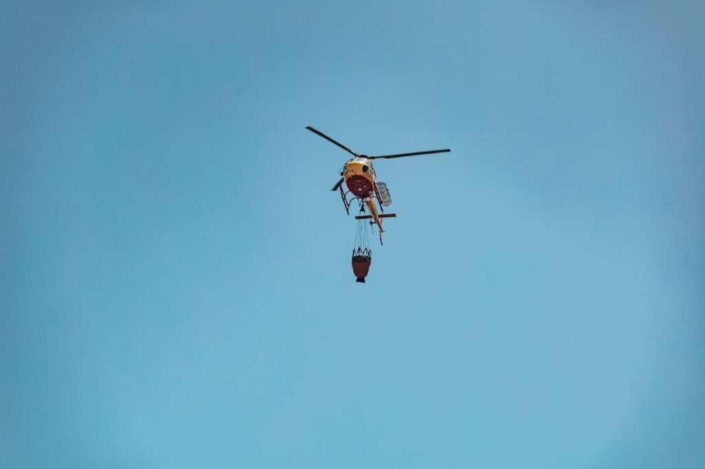 A helicopter equipped for firefighting carries a water bucket against a clear blue sky.